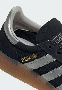 Baskets Adidas Spezial noires avec des bandes argentées, le texte "SPEZIAL" en doré, une languette de talon argentée et une semelle externe translucide couleur gomme.