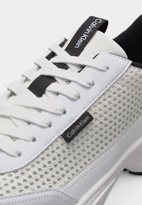 Λευκό αθλητικό παπούτσι Calvin Klein με λευκά κορδόνια, μαύρη γλώσσα με το λογότυπο και πλέγμα στην πλευρική επιφάνεια που δείχνει υφή.