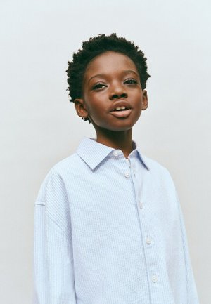 Copil negru tânăr cu păr scurt și creț, purtând o cămașă cu dungi bleu deschis și nasturi, privind înainte pe un fundal neutru.