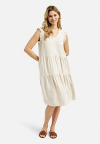 Beiges, gestuftes Kleid mit V-Ausschnitt und Rüschensäumen. Leichter Stoff fällt locker und wird mit braunen Slippers kombiniert.