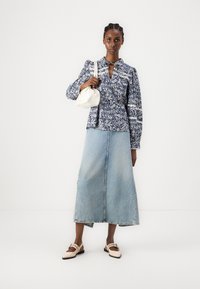 Bluse med blomsterdesign, lange ermer i marineblå og hvit, kombinert med en lys blå denimmaxiskjørt og lysebeige leppesko. Holder en liten hvit veske.