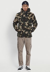 Chaqueta de forro polar con patrón de camuflaje, capucha, cierre de cremallera y dos bolsillos frontales, combinada con pantalones cargo claros y zapatillas en blanco y negro.