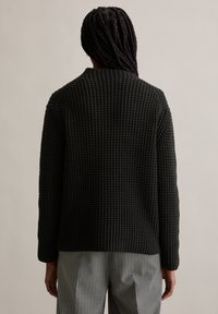 Schwarzer Strickpullover mit strukturiertem Muster, runder Halsauschnitt und langen Ärmeln, getragen zu grauen gepinnten Hosen.