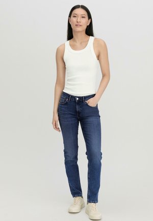 Femme debout, main dans la poche, portant un haut blanc sans manches, un jean bleu et des baskets blanches sur un fond clair uni.