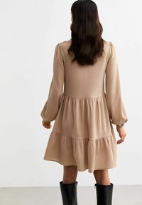 Beige tiered dress with a textured fabric, long puff sleeves, and an elasticated waist. Worn with knee-high black boots.