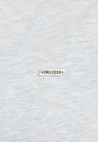 Un tejido de camiseta gris con una textura suave presenta una pequeña etiqueta metálica con la inscripción "SMILODOX" fijada en la parte frontal.