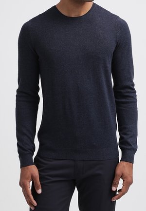 Mann trägt einen dunkelblauen, enganliegenden Pullover mit Rundhalsausschnitt und langen Ärmeln sowie schwarze Hose, steht mit entspannt an den Seiten hängenden Armen.