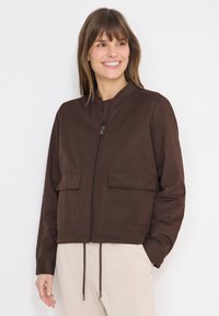 Femme souriante portant une veste zippée marron avec poches avant et un pantalon beige, debout devant un fond blanc.