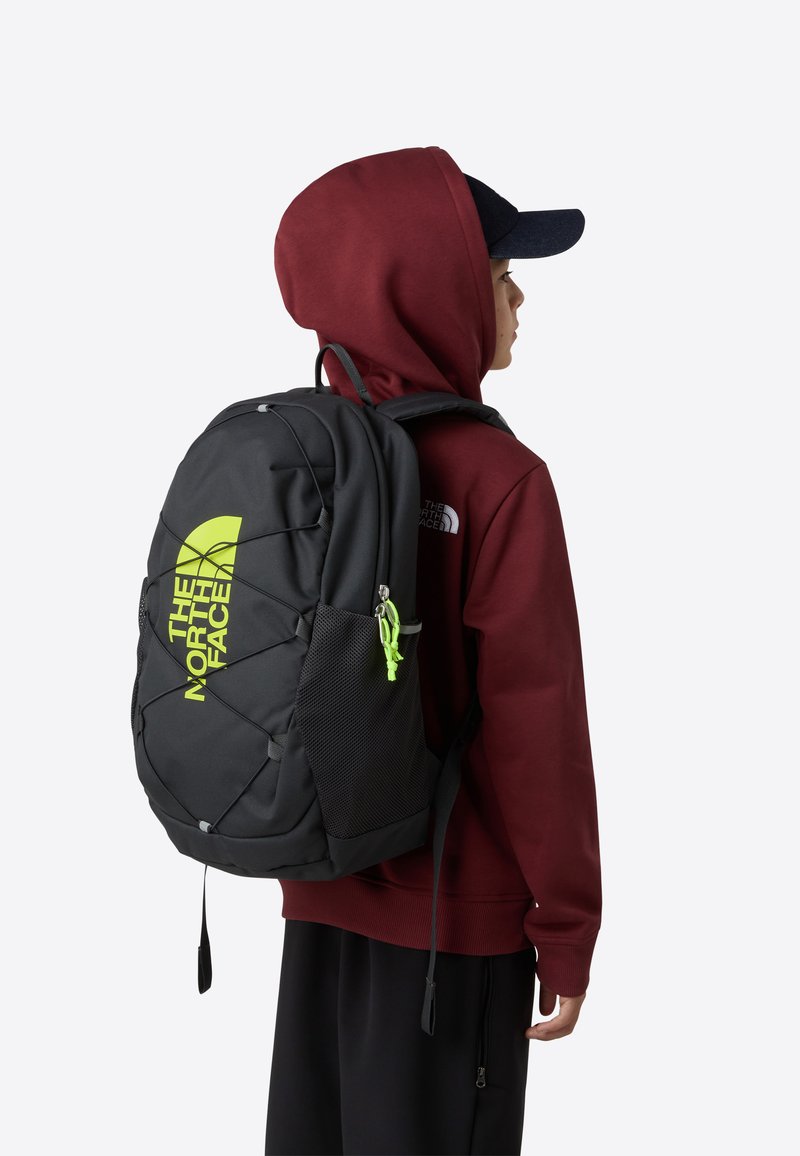 Svart ryggsekk med justerbare stropper, nettinglommer på sidene, neongrønn logo og glidelåsdetaljer. Bæres av en person i en burgunder hettegenser og caps.