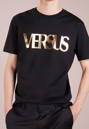 Personne portant un t-shirt noir avec un grand texte métallique doré "VERSUS" sur la poitrine, mains dans les poches.