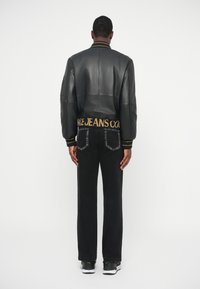 Άνδρας που φοράει μαύρο δερμάτινο μπουφάν και μαύρο τζιν, γυρισμένος προς τα πίσω σε λευκό φόντο.