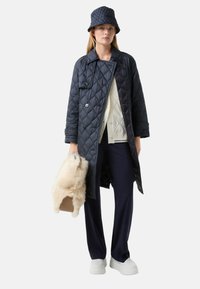 Manteau matelassé navy avec taille ceinturée, associé à un chapeau seau coordonné, un pull crème et un sac à main beige en peluche. Chaussures blanches à plateforme.