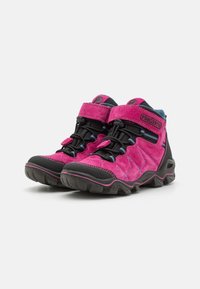 Wysokie buty trekkingowe mają jasnoróżową zamszową cholewkę z czarnymi akcentami, teksturowaną gumową podeszwę oraz niebieskie detale. Zapięcie na regulowany pasek.