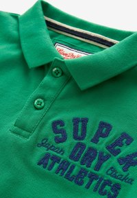 Zelená polo košeľa vyrobená z textúrovaného bavlneného materiálu. Obsahuje golier, dva zelené gombíky a na prednej strane je vyšitý námornícky logo "SUPER DRY ATHLETICS".
