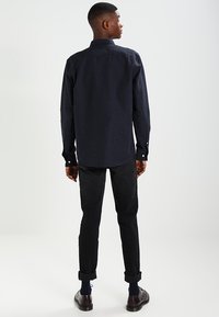 Camicia blu navy con motivo a macchie, maniche lunghe e polsini con bottoni. Indossata con pantaloni neri arrotolati e scarpe scure. Sfondo semplice.