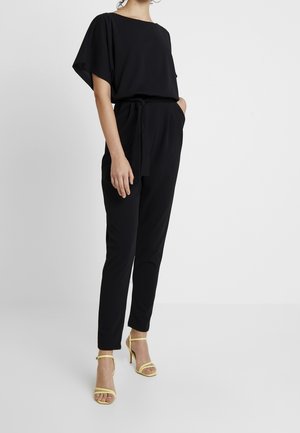 Kvinde iført en sort kortærmet jumpsuit med taljebånd og gule høje hæle sandaler, der står mod en ensfarvet lys baggrund.