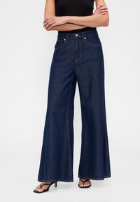 Široke kavbojke iz temno modrega jeansa z visokim pasom, sprednjimi žepi in šivnimi detajli. Kombinirane s črnimi čevlji z odprtimi prsti.