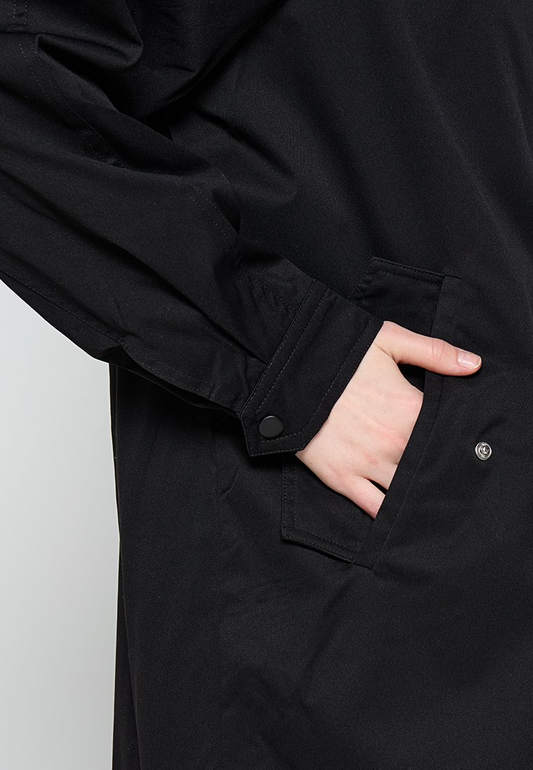 Hand teilweise in der vorderen Tasche einer schwarzen Langarmjacke mit Knopfmanschette und Druckknopfverschluss.