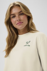 Felpa color crema con un logo verde "racket club" e una grafica di racchette incrociate sul lato sinistro del petto, realizzata in un tessuto morbido.
