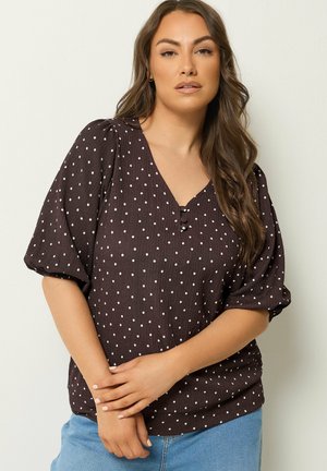 Kvinde iført en mørkebrun kortærmet bluse med hvide prikker og lyseblå jeans, står med hænderne foldet foran sig.