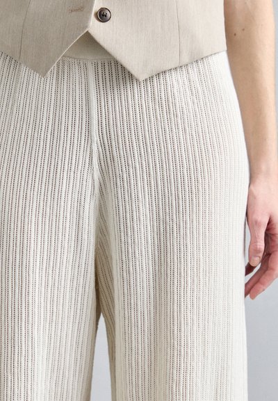 Beige geribbelde gebreide broek met wijde pijpen en een textuurafwerking. Kenmerken zijn een hoge taille en rechte snit, gecombineerd met een linnen vest.