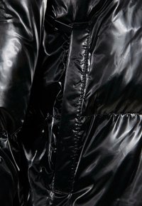 Veste matelassée noire en tissu brillant et texturé. Dotée de détails de couture, d'une sangle verticale et de fronces à l'ourlet pour lui donner de la forme.