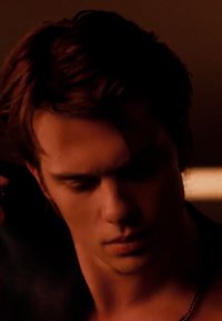 Jeune homme aux cheveux bruns regardant vers le bas dans une lumière chaude et tamisée, affichant une expression calme ou introspective.
