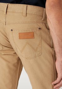 Pantalones de sarga de algodón en color tan que presentan una etiqueta de cuero de Wrangler, dos bolsillos traseros con detalles de costura y un diseño de pierna recta.