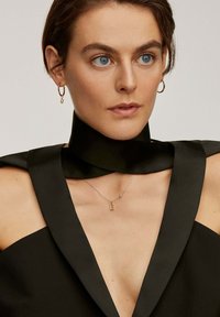 Vestido de satén negro con solapas anchas y un escote profundo, adornado con un collar plateado con un colgante en forma de 'E' y pequeñas embellishments, acompañado de pendientes de aro dorados.