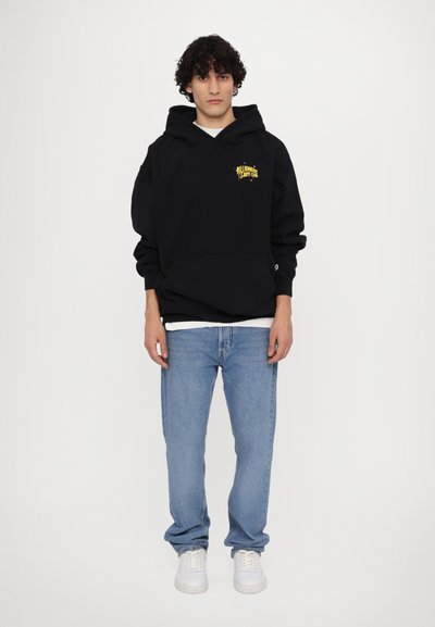 Haut à capuche noir avec une poche avant et un logo jaune, associé à un jean en denim bleu clair et des baskets blanches. Le modèle se tient devant un fond blanc.