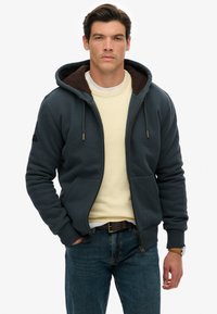 Man in een donkerblauwe ritshoodie met een bruine voering, een crèmekleurige trui, blauwe jeans en een horloge, staand met één hand in de zak.