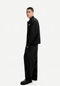 Black lightweight jacket with a relaxed fit, featuring an embroidered logo. Paired with black loose-fitting pants with button details and textured fabric.
