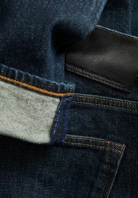 Jean en denim bleu foncé en gros plan montrant un revers plié avec intérieur gris clair et un patch de marque en cuir noir aux bords cousus.
