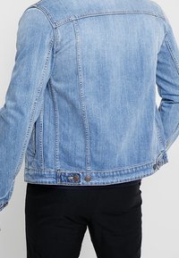 Giacca di jeans blu chiaro con un design classico, presenta due bottoni in vita e cuciture visibili lungo le cuciture e le spalle.