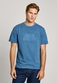 Mėlynas medvilninis marškinėliai trumpomis rankovėmis su iškilniu užrašu "PEPE JEANS" priekyje. Apvalus iškirpimas ir tekstūruota medžiaga, derinama su tamsiais džinsais.