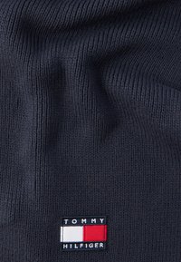 Σκούρο μπλε πλεκτό ύφασμα με μικρό ορθογώνιο ταμπελάκι λογότυπου Tommy Hilfiger που διαθέτει κόκκινα, λευκά και ναυτικά μπλε χρώματα.