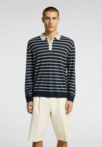 Polo a righe blu navy e bianche con maniche lunghe e colletto beige, abbinato a pantaloni corti di un leggero color crema. Tessuto morbido, vestibilità comoda.