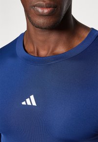 Sötétkék sportpóló lekerekített nyakkal, bal mellején három átlós csíkkal díszített fehér logóval. Simaság jellemzi.