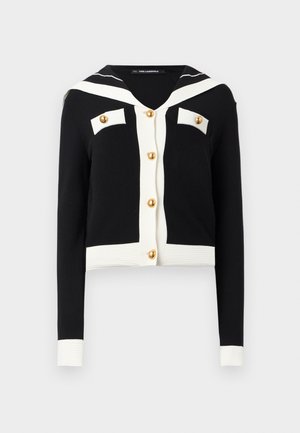 Μαύρη ζακέτα με λευκό γιακα σε στυλ ναυτικού, λευκό περιθώριο στα μανίκια και στην κάτω άκρη, χρυσά κουμπιά μπροστά και σε δύο τσέπες στο στήθος.