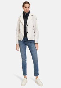 Light cream double-breasted jacket with a structured collar, layered over a black quilted jacket. Worn with fitted blue jeans and white sneakers.