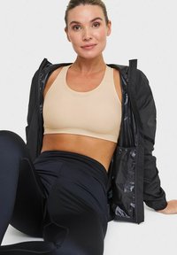 Svart lättviktsjacka över en beige sport-bh och svarta högmidjade leggings, alla tillverkade av släta, töjbara material.