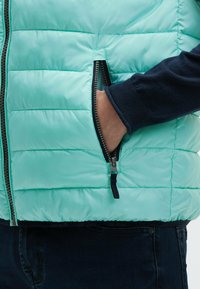 Mano nella tasca di una giacca trapuntata blu chiaro con zip, indossata sopra un maglione blu scuro e jeans scuri.