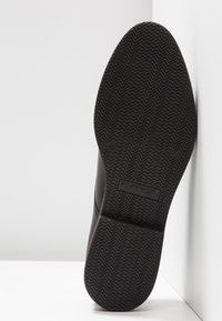 Semelle de chaussure en caoutchouc noir avec motif de bande de roulement en zigzag et logo de marque embossé. Bords lisses et design minimaliste. Orientation verticale.