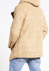 Giubbotto puffer beige con cappuccio, caratterizzato da un design trapuntato, texture liscia e tasche laterali con zip, abbinato a jeans in denim azzurro chiaro.