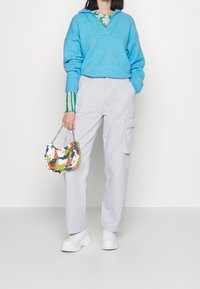 Hellblauer Strickpullover mit V-Ausschnitt, kombiniert mit hellgrauen Cargohosen. Die Tasche ist mit bunten Pailletten verziert, und weiße Turnschuhe runden den Look ab.