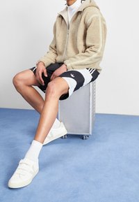 Tan fluffig zip-up jacka med vita ränder, svart- och vitrandiga shorts, vita sneakers med kardborreband, sittande på en metallbox.