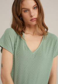 Světl zelený top s krátkým rukávem a výstřihem do V, s texturovaným vzorem v chevron stylu a volným střihem.