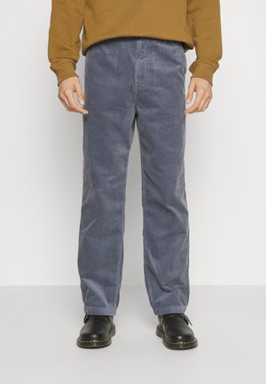 Mann trägt blaue Cordhosen, schwarze Stiefel und ein braunes Langarmhemd, steht vor einem schlichten Hintergrund.