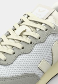 Sneaker grigio chiaro con tomaia in rete testurizzata, accenti in suede chiaro, lacci crema e un'etichetta bianca con un "V" sulla linguetta.