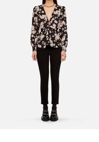 Vrouw in een zwarte bloemenblouse met lange mouwen en een gedraaide voorkant, zwarte slim-fit broek, zwarte gespt loafers en een paarse halsketting.